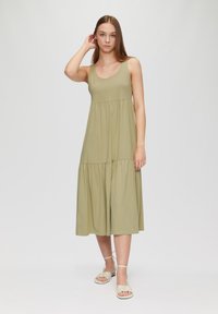 Robe sans manches vert olive avec un décolleté en forme de rond et un design à volants, offrant une silhouette fluide. Assortie à des sandales à lanières de couleur claire.