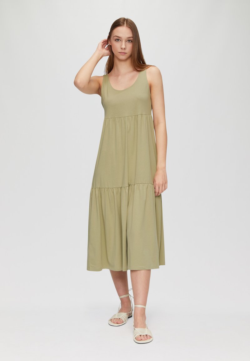 Robe sans manches vert olive avec un décolleté en forme de rond et un design à volants, offrant une silhouette fluide. Assortie à des sandales à lanières de couleur claire.