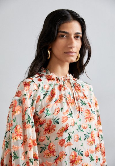Blusa floral con fondo blanco roto y grandes estampados de amapolas naranjas. Escote con volantes y detalle de cordón, y mangas sueltas.