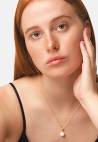 Collana in oro con pendente a forma di perla bianca tonda, indossata sulla pelle nuda con un top nero; design semplice ed elegante.