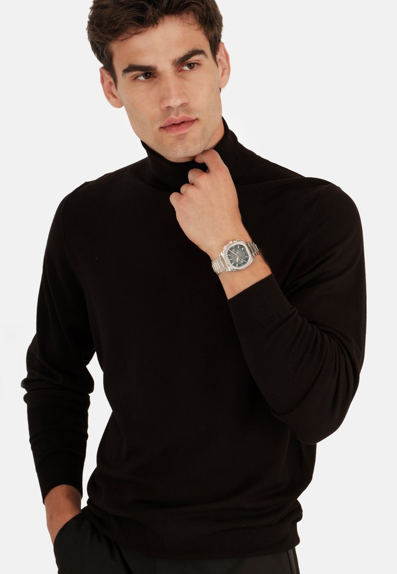 Un pull col roulé noir en tissu lisse, avec des poignets côtelés et une coupe ajustée. Une montre en argent avec un cadran gris est portée.