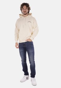 Felpa crema con cappuccio e lacci, tasca frontale; abbinata a jeans blu scuro e sneaker bianche; il modello sta in piedi con le mani in tasca.