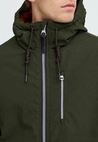 Giacca a zip argentata con cappuccio verde oliva, dettagli neri e un laccetto testurizzato. Materiale liscio con una vestibilità strutturata.