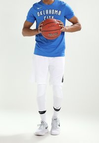 Niebieska koszulka Nike z nadrukiem "OKLAHOMA CITY", białe spodenki, białe legginsy do kolan oraz białe sneakersy Nike, trzymająca piłkę do koszykówki.