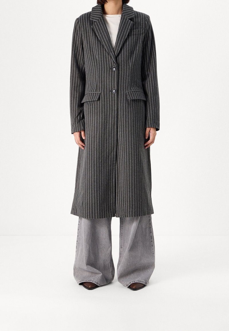 Šedý pruhovaný dlouhý kabát s klopovými límci a dvěma předními kapsami. Materiál má hladkou texturu, v kombinaci se širokými šedými džíny a špičatými botami.