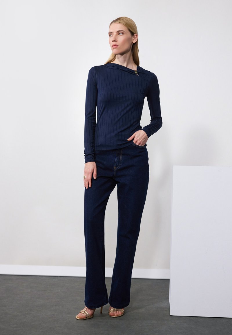 Top a maniche lunghe a coste navy con scollatura asimmetrica, abbinato a jeans blu scuro a gamba dritta e sandali leggeri con cinturini.