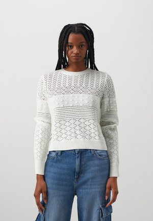 White knitted sweater featuring a mix of open and closed patterns, ribbed cuffs, and a rounded neckline. Paired with blue denim.