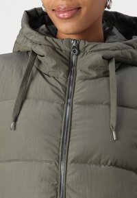 Veste matelassée vert olive avec fermeture éclair à l'avant, capuche et cordons de serrage. Fabriquée en tissu léger et brillant, avec des fermetures éclair en métal argenté.