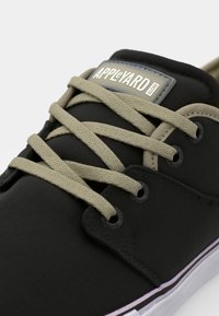 Black sneaker with textured fabric, beige laces, and a rubber sole. Features a brand label on the tongue and circular eyelets.