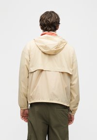 Άνδρας με μπεζ μπουφάν με κουκούλα και ελαστικά μανίκια, που φοράει χακί παντελόνι, φωτογραφημένος από πίσω μπροστά σε απλό λευκό φόντο.