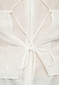 Λευκό πουκάμισο με πίσω σχέδιο από σταυρωτές τιράντες και δεσίματα στη μέση, κατασκευασμένο από ελαφρύ, υφασμένο ύφασμα.