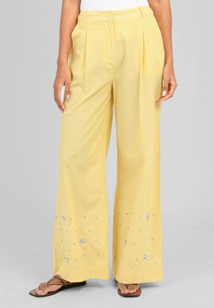 Pantalon jaune taille haute à jambes larges avec de fines rayures blanches et de petites décorations florales près de l'ourlet, porté avec des tongs marron.