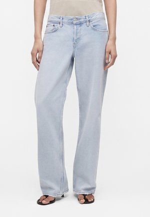 Person iført lyseblå bukser med vide ben og sorte åbne sandaler, der står mod en ensartet lys baggrund.