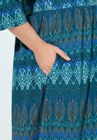 Teal mekko, jossa on geometrinen kuosi sinisen, vihreän ja ruskean sävyissä. Vyötäröllä sauma ja sivutasku. Pehmeä kangas.