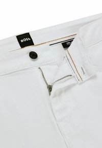 Pantaloni in denim bianco con zip argentata parzialmente aperta, bottone nero e etichetta nera con il testo "BOSS" sulla vita.