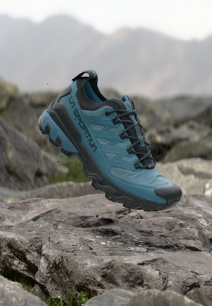 Chaussure de trail running La Sportiva bleue et noire en l’air au-dessus d’un terrain rocheux extérieur avec un arrière-plan montagneux flou.