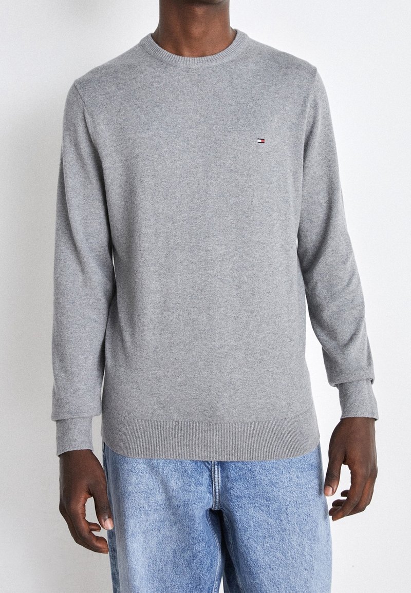 Homme portant un pull gris clair à col rond avec un petit logo Tommy Hilfiger sur la poitrine et un jean bleu clair, debout devant un fond blanc.
