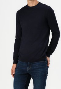 Person som bär en figursydd svart långärmad tröja med rundad hals och mörkblå jeans, står med ena handen i fickan mot en vit bakgrund.