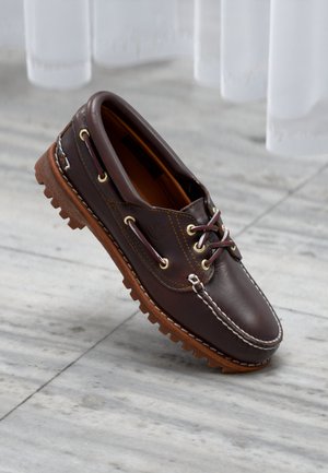 Brun læder bådsko med rundet tå, hvide syninger, guldøjer og en brun gummisål med mønstre til trækkraft.