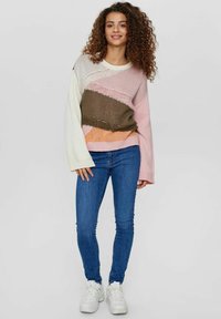 Šareno pleteni pulover s krem, ružičastim, maslinastim i narančastim dijelovima. Labav dizajn s širokim rukavima. Uparen s plavim uskim trapericama.