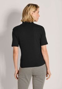 Frau mit kurzem blonden Haar, die ein schwarzes kurzärmliges Oberteil und Hosen mit Schwarz-Weiß-Hahnentrittmuster trägt und von der Kamera abgewandt ist.