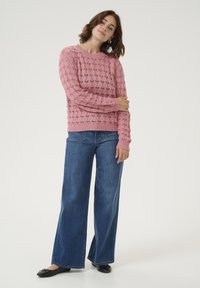 Maglione rosa lavorato a maglia con un motivo testurizzato, abbinato a jeans blu a gamba larga e ballerine nere. Il maglione ha un collo a giro e polsini a coste.