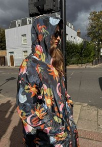 Must mustriga vihmakeep erksate värvidega nagu oranž ja roosa, kapuutsi ja küljetaskutega, valmistatud kerge ja läikiva materjaliga.