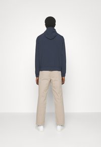 Hanorac cu glugă de culoare bleumarin, cu o croială elegantă, asortat cu pantaloni bej dreți și adidași albi. Textură netedă, design minimalist.