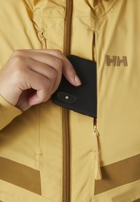 Gelbe Jacke mit einer Reißverschlusstasche; eine Hand steckt ein schwarzes Smartphone hinein. Mit braunen Akzenten und einem Logo auf der Brust. Strukturierten Stoff.