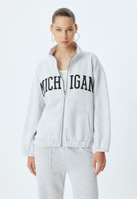 Chaqueta gris con cremallera hecha de una tela suave, con la inscripción "MICHIGAN" en negro en el pecho, que cuenta con puños elásticos y bolsillos laterales.