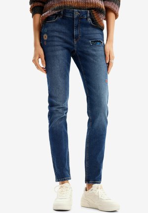 Kobieta ubrana w niebieskie dżinsy z kolorowymi, haftowanymi wzorami słońca, z przetarciem na udzie, zestawione z białymi trampkami.