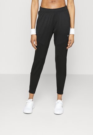 Atletisch persoon draagt zwarte taps toelopende Nike sportbroek, witte polsbandjes en witte sneakers, staand tegen een eenvoudige lichte achtergrond.