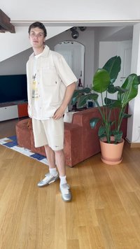 Camisa de botones de manga corta color crema, camiseta gráfica debajo, pantalones cortos beige con textura suave y zapatillas plateadas, con una planta y un sofá marrón al fondo.
