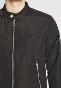 Svart bomberjacka i slät nylon med hög krage, framdragkedja och sidofickor. Har en diskret logotyp på bröstet.