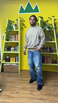 Hanorac bej, blugi albaștri și pantofi negri purtați de o persoană într-o cameră cu pereți galbeni și rafturi verzi pentru cărți. Plantele sunt vizibile.