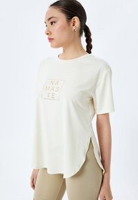 Cremefarvet kortærmet t-shirt med et firkantet "NAMASTE" grafisk motiv i guld, med sideslidser og en afslappet pasform. Glat, letvægtigt stof.