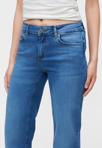 Midtdelen av en person som har på seg blå denim-jeans og en cropped hvit topp, stående med armene avslappet ned langs sidene.