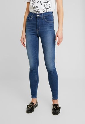 Vaqueros ajustados de color azul, combinados con mocasines negros que tienen detalles en hebilla dorada y una camiseta blanca con gráficos.