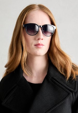 Occhiali da sole neri e grigi a forma di occhio di gatto, con lenti sfumate e dettagli lucidi, indossati con un cappotto nero e un collo semplice.