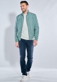 Helle türkisfarbene Bomberjacke mit Stehkragen, Vorderreißverschluss und zwei Seitentaschen, kombiniert mit einem hellen Pullover und dunklen Jeans.