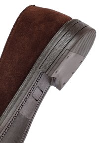 Suola di scarpa in suede marrone con tacco in gomma testurizzata e dettagli cuciti sul lato, mostrata in una vista ravvicinata angolata.
