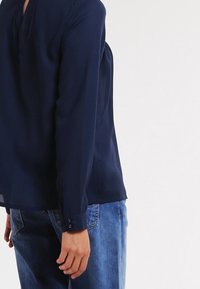 Maglietta a maniche lunghe blu navy con placca frontale e polsini con bottone, realizzata in tessuto leggero. Indossata con jeans azzurri.