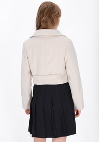 Cremfarbener, verkürzter Blazer mit breitem Kragen und zwei Knöpfen in der Taille. Kombiniert mit einem plissierten schwarzen Rock.
