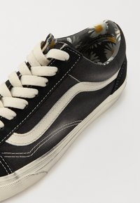 Baskets noires en toile avec des accents blancs, surface texturée et lacets beige. L'intérieur présente un motif floral en blanc et jaune.