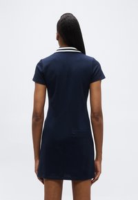 Γυναίκα με μακριά σκουρόχρωμα μαλλιά, φοράει μπλε σκούρο φόρεμα με κοντά μανίκια και γιακά με λευκές ρίγες, γυρισμένη πλάτη σε απλό φόντο.