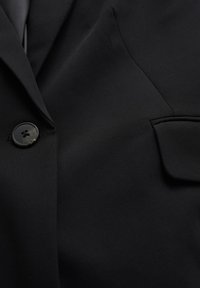 Černý blazer z hladkého materiálu, s jedním knoflíkem, klopovým límcem a kapsou na prsou, ukazující na přizpůsobený design.
