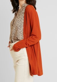 Kvinna i en rostorange kofta över en beige leopardmönstrad blus som är instoppad i högmidjade off-white byxor med händerna i fickorna.