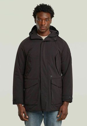 Homme portant un manteau d'hiver noir à capuche avec de grandes poches avant et un jean bleu, debout devant un fond clair uni.