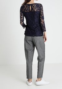 Žena stojící, oblečená v námořnickém modrém krajkovém topu s dlouhými rukávy, šedých upravených kalhotách a bílých teniskách s růžovým detailem na podpatku, čelem otočená pryč.