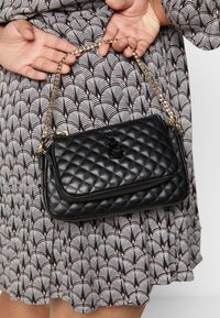 Hænder holder en sort quiltet taske med guldkæde imod en mønstret kjole med bølgende design.